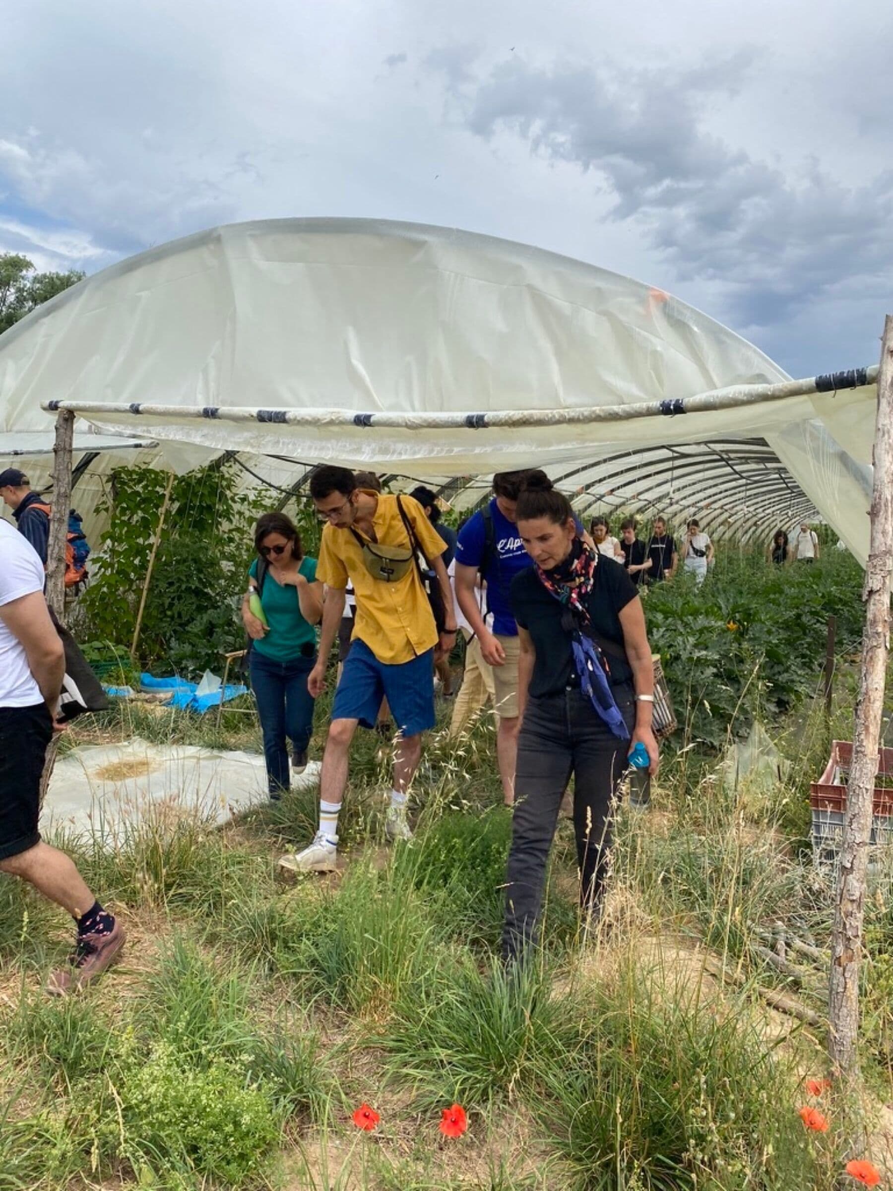 A group of people walks through a grassy area under a large, arched greenhouse structure. The sky is cloudy, creating a serious and focused atmosphere.